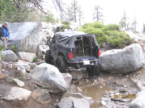 1994 Ford Bronco - This modified SUV is navigating a challenging off-road trail with large rocks and muddy puddles.