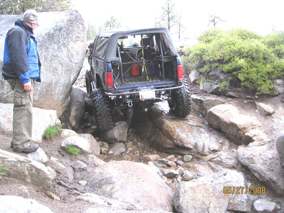 Dark Bronco with roll cage and mud tires crawling over wet granite rocks, spotter watching