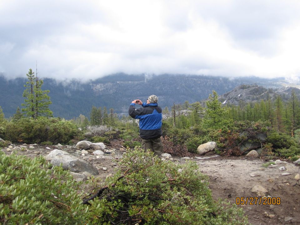 Person in blue jacket photographing a cloudy mountain ridge from a dirt trail