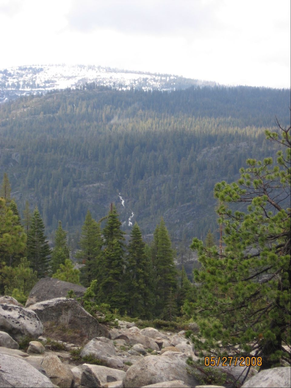 Mountain overlook with granite boulders, pine forest, waterfall and snowy ridge