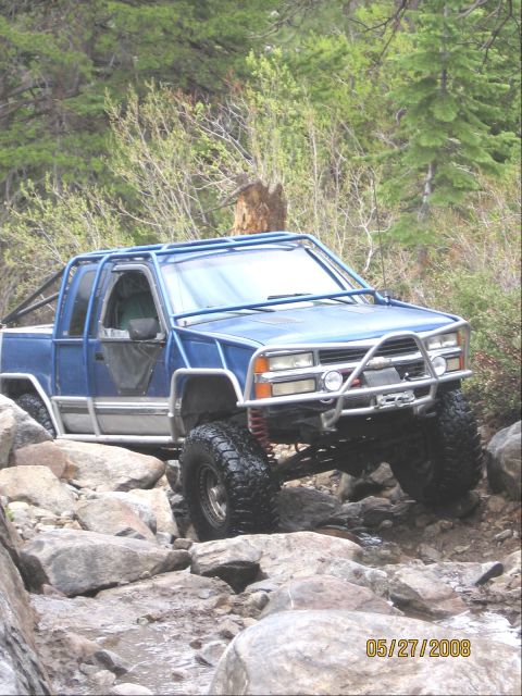 1994 Ford Bronco - A blue pickup truck with a custom roll cage is shown off-roading over large rocks on a wooded trail.