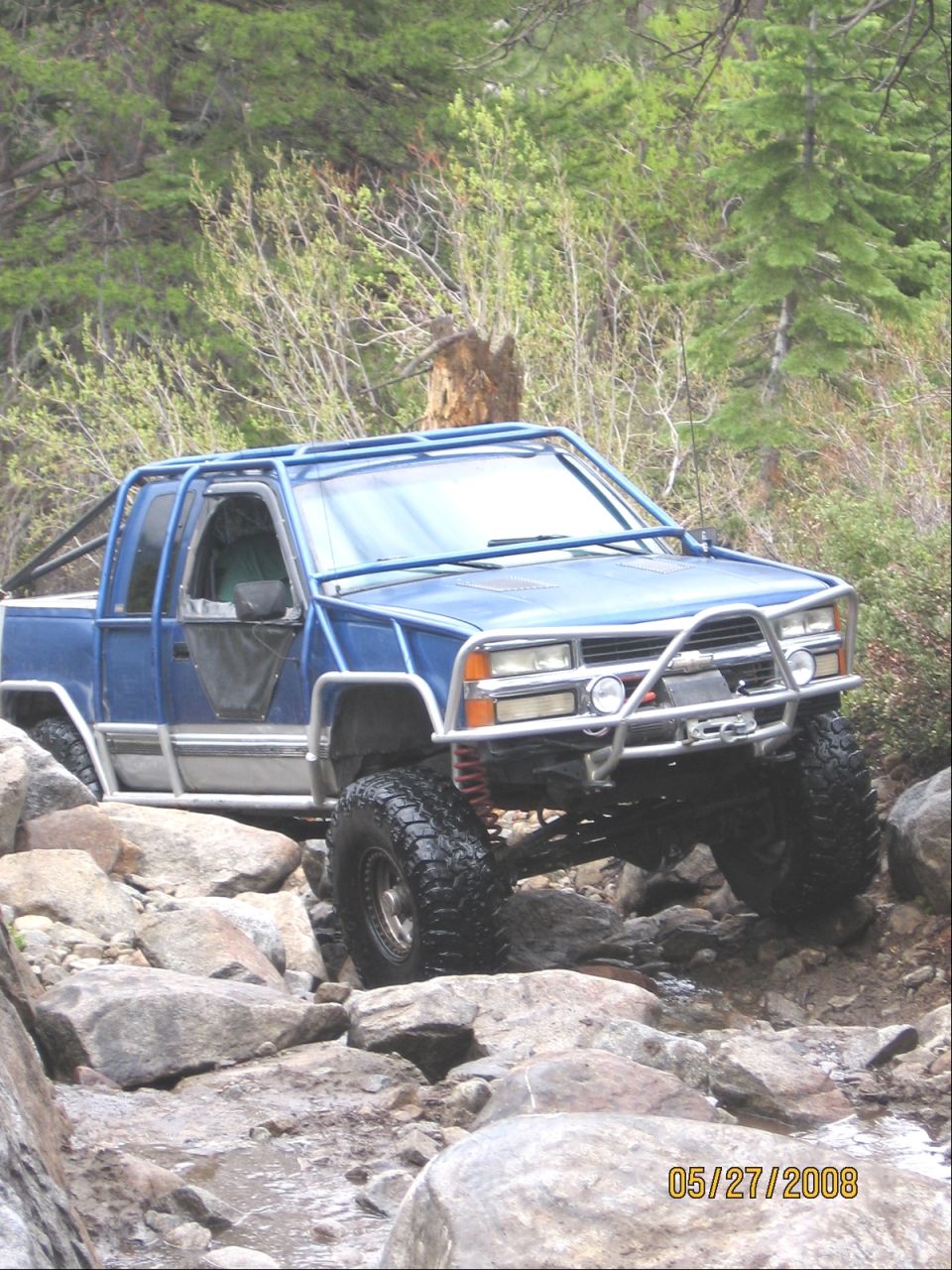 Blue 1990s Chevy pickup with exterior roll cage and winch crawling a rocky creek bed