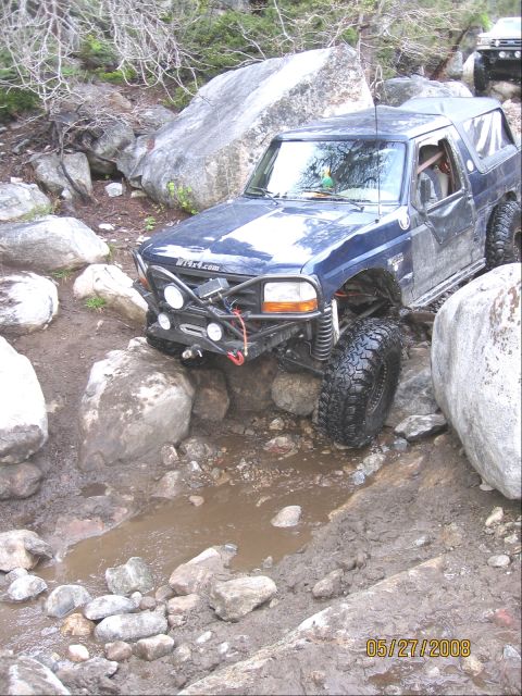 1994 Ford Bronco - This modified SUV is tackling a challenging off-road trail with large rocks and water obstacles.