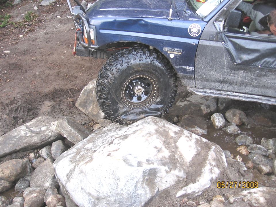 Dark blue Ford Bronco with winch and large mud tire crawling over boulders on muddy trail