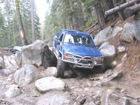 1994 Ford Bronco - A blue off-road vehicle maneuvers through a challenging rocky terrain in a forest.