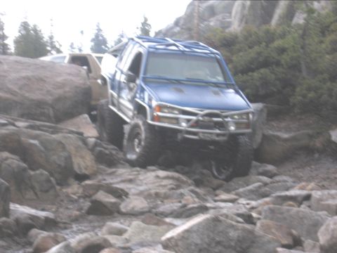 1994 Ford Bronco - A blue SUV with an external roll cage is shown navigating a challenging rocky trail, with another vehicle partially visible behind it.