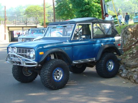 1994 Ford Bronco - Modified blue Ford Bronco with large tires and soft top, parked on pavement near a rocky incline.