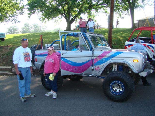 Silver Ford Bronco with pink and blue stripes, roll cage, and off-road tires. Two people stand beside it