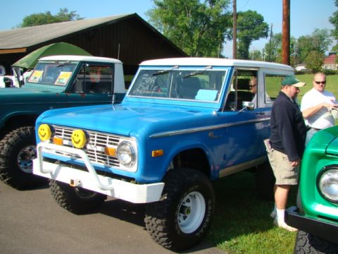 1994 Ford Bronco - A blue Ford Bronco with a white roof and bumper is parked next to a green truck, with a "FOR SALE" sign visible on its windshield.