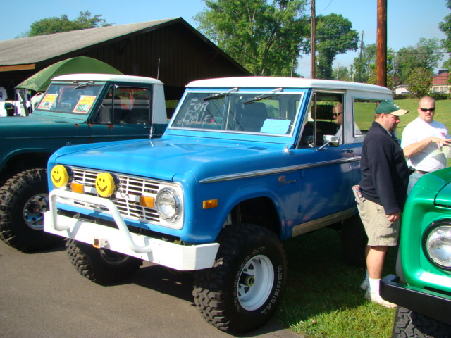 Blue Ford Bronco with white roof and bumper, "FOR SALE" sign on windshield, parked next to green truck