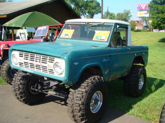 Teal Ford Bronco with lifted suspension and large tires. Two yellow signs on windshield