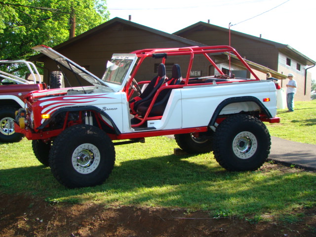 White and red modified Ford Bronco with roll cage and large tires. Another Bronco is partially visible to the left