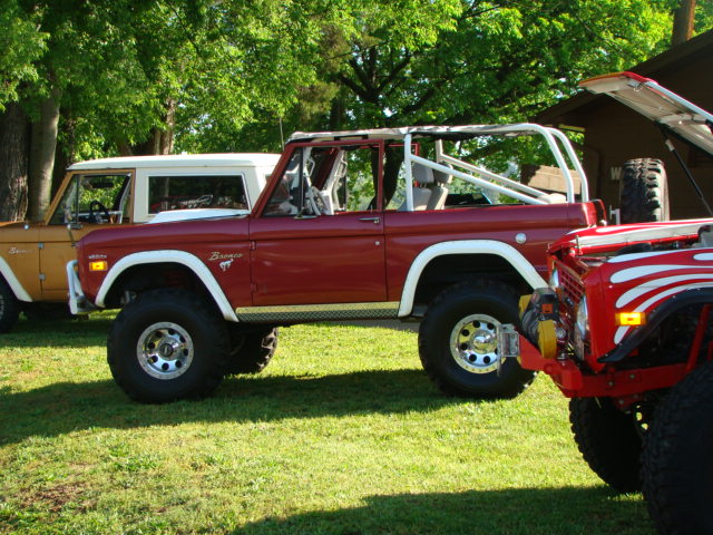 Red Ford Bronco with white roll cage parked next to a tan Bronco. Another red Bronco is partially visible in the foreground