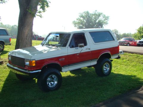 1994 Ford Bronco - A two-tone red and white Ford Bronco is parked on a grassy area next to a tree, with other vehicles visible in the background.