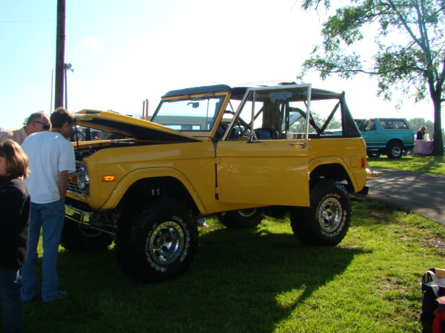 Yellow 1978 Ford Bronco with hood open, parked on grass. Another blue SUV is visible in the background