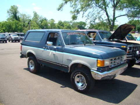 1994 Ford Bronco - A two-tone Ford Bronco is parked in a lot with other vehicles, including a truck with an open hood.