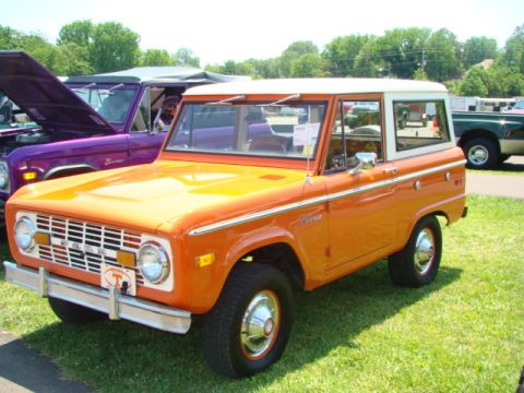1994 Ford Bronco - Orange and white Ford Bronco parked on grass with other vehicles in the background.