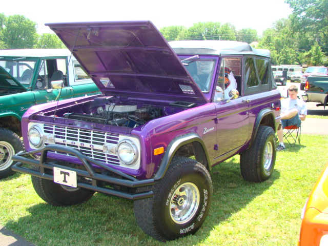 Purple 1978 Ford Bronco with hood open, parked on grass next to a green Bronco