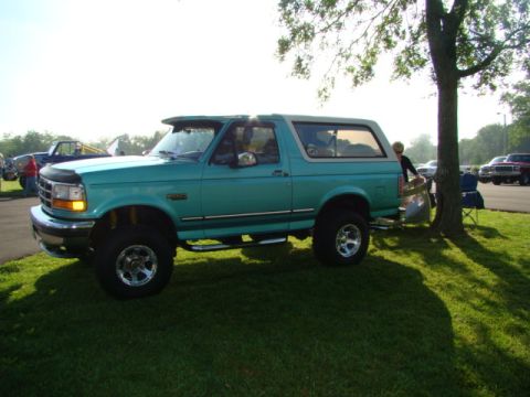 1994 Ford Bronco - A teal Ford Bronco is parked on a grassy area next to a tree, with other vehicles visible in the background.