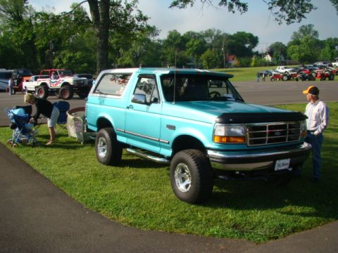1994 Ford Bronco - A turquoise 1994 Ford Bronco with a white top is displayed at an outdoor event, with other vehicles visible in the background.