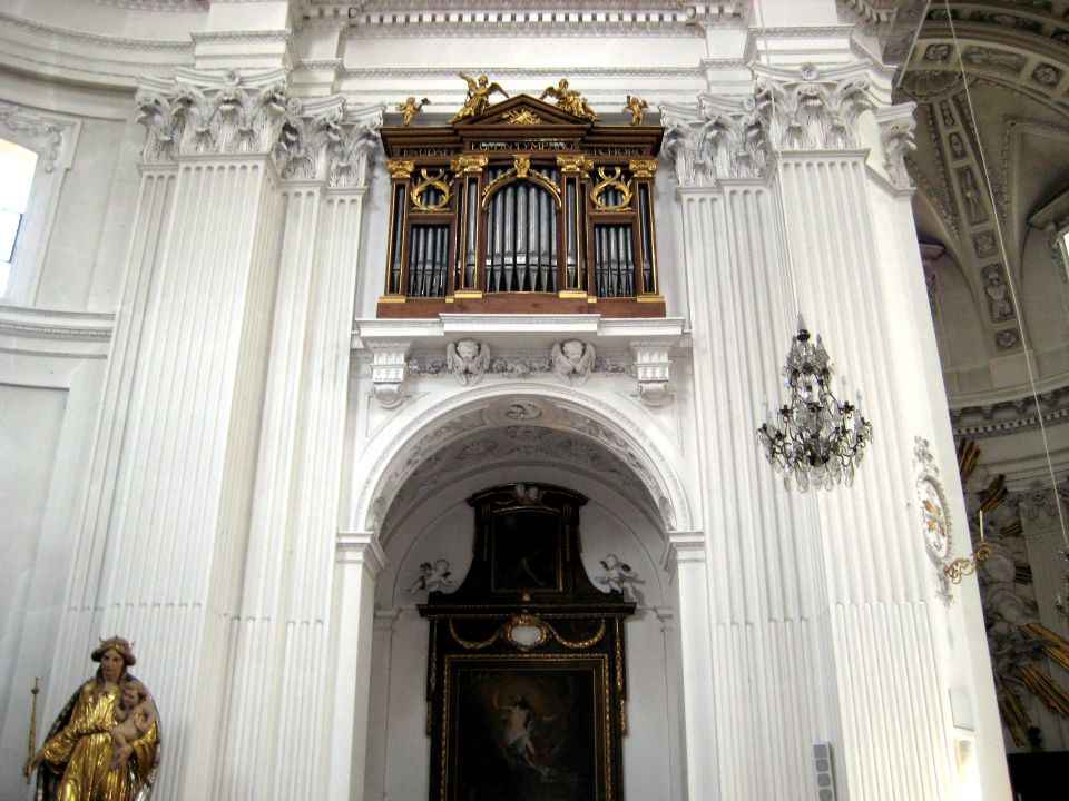 Interior of a church with a large pipe organ mounted high on the wall above a painting. A statue is visible on the left