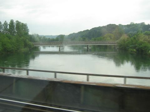 1996 Ford Bronco - A bridge spans a calm river, bordered by lush green trees and rolling hills under an overcast sky.