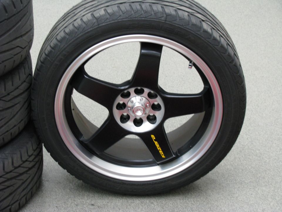 Black five-spoke aftermarket wheel with silver lip, yellow Kazera logo, mounted on a tire. Stack of tires visible on left