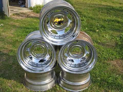 1981 Ford Bronco - Four polished aluminum three-piece wheels stacked in a pyramid on grass outside a shed, showing deep-dish outer barrels with visible assemb…