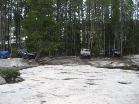 1989 Ford Bronco - Multiple off-road vehicles are parked in a clearing surrounded by tall pine trees, with a rocky area in the foreground.