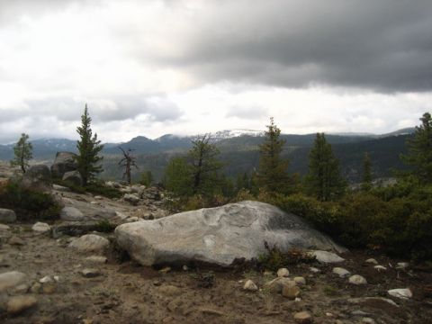 1989 Ford Bronco - A scenic view of a mountainous terrain with scattered rocks and evergreen trees, under a dramatic cloudy sky.