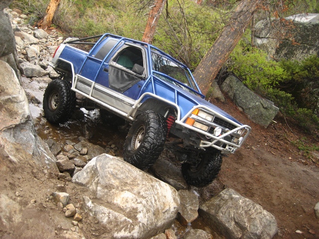 Blue compact 4x4 with external roll cage crawling over boulders by a creek