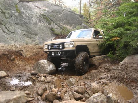 1989 Ford Bronco - A lifted Ford Bronco navigates a rocky, off-road trail with dense trees and large boulders.