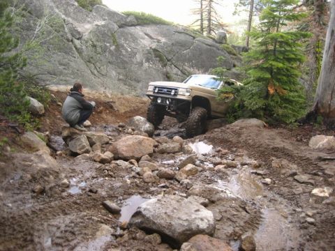1989 Ford Bronco - An off-road SUV tackles a challenging rocky and muddy terrain on a wooded mountainside.