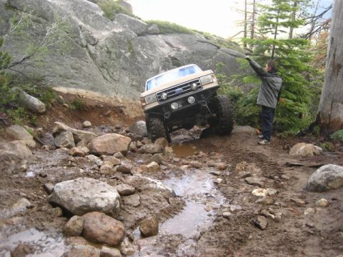 1989 Ford Bronco - A tan 1987 Ford Bronco tackles a challenging off-road path filled with mud and rocks, while a bystander observes.