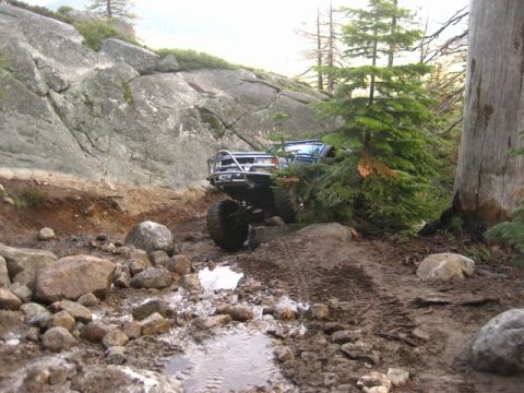 1989 Ford Bronco - A modified off-road vehicle tackles a challenging, muddy, and rocky trail in a wooded area.