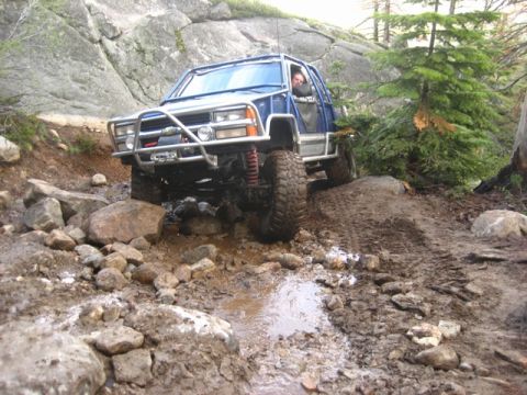 1989 Ford Bronco - A modified blue SUV tackles a challenging off-road course with large rocks and mud.
