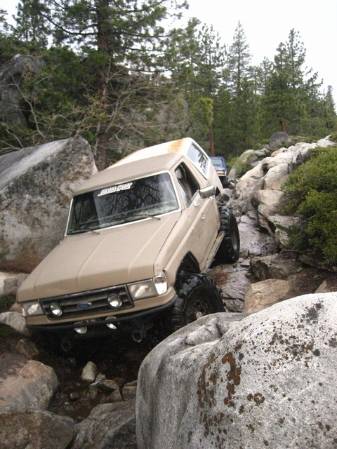 1989 Ford Bronco - A tan Ford Bronco tackles a challenging rocky off-road path, with another vehicle following in the distance.