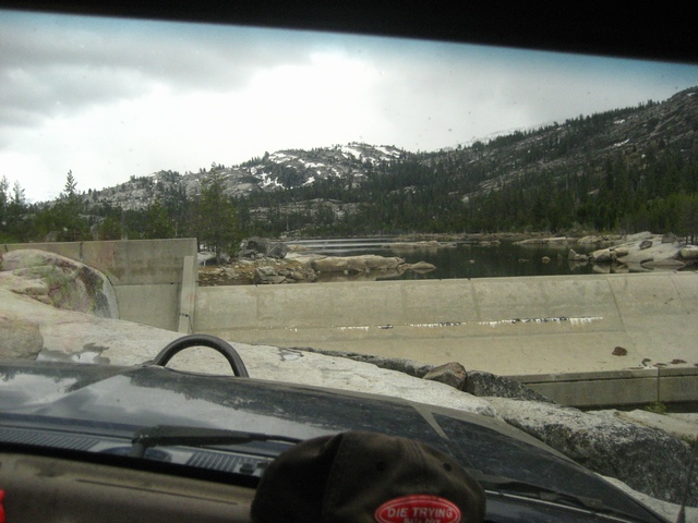 Through-windshield view over the hood of a concrete dam and granite mountain reservoir