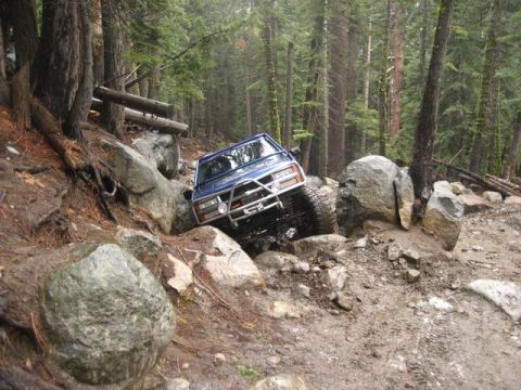 1989 Ford Bronco - A blue SUV is carefully maneuvering over large rocks on a steep, wooded trail.