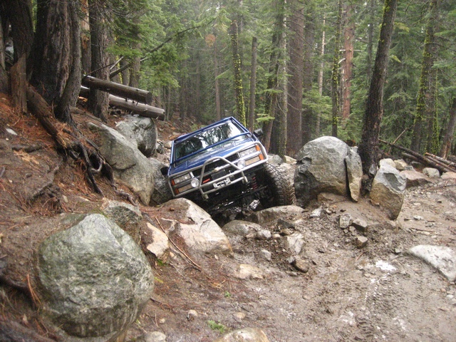 Blue 4x4 with brush guard crawling nose-down over boulders on a forest trail