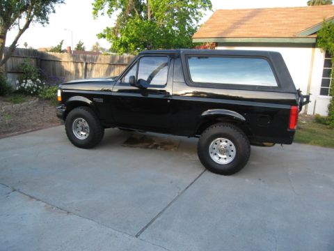 1986 Ford Bronco - A black two-door Ford Bronco is parked on a concrete driveway in front of a house and a wooden fence. The vehicle has aftermarket wheels an…