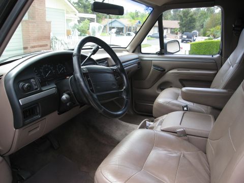 1986 Ford Bronco - The interior of a tan Ford Bronco is shown, featuring leather seats, a steering wheel, and the dashboard with gauges. The view looks out th…
