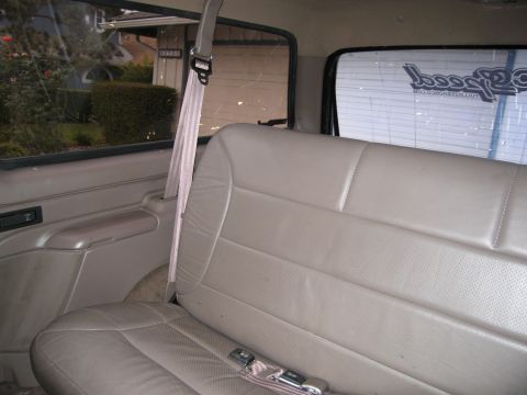 1986 Ford Bronco - The interior of a vehicle shows a tan leather bench seat with a pink seatbelt. The view looks towards the rear of the vehicle, with a windo…