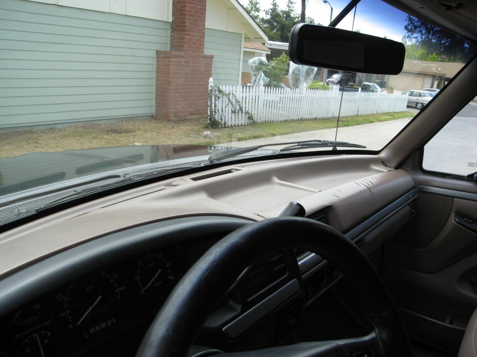 Interior view of a vehicle dashboard and steering wheel, looking out windshield at house and fence.