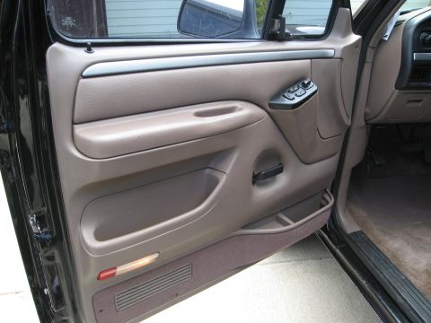 1986 Ford Bronco - The interior of a vehicle's driver side door panel is shown, featuring a brown textured armrest, a black pull handle, and a window control…