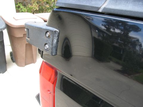 1986 Ford Bronco - A close-up view of the rear corner of a black vehicle, showing a black metal bracket with two bolts attached to the body. A red taillight i…