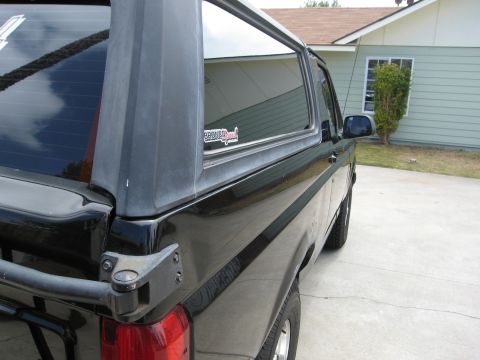 1986 Ford Bronco - The rear passenger side of a black Ford Bronco is shown parked on a concrete driveway in front of a light green house. A sticker reading "B…
