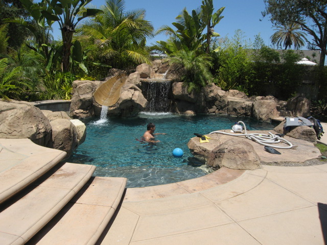 A man swims in a backyard pool with a water slide and rock waterfall.