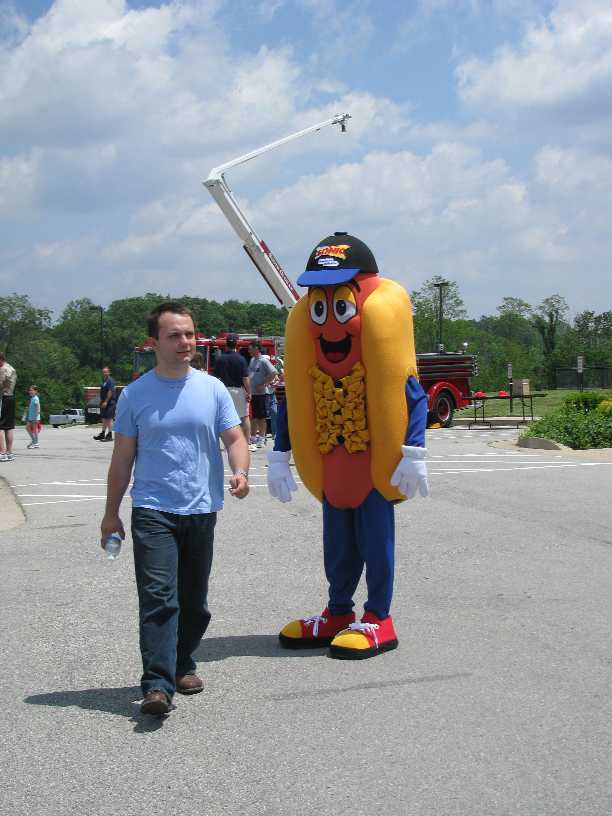 A man walks next to a hot dog mascot with a cherry picker and fire truck in the background.