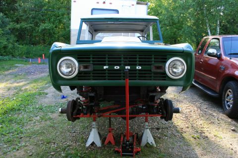 1974 Ford Bronco - A green 1974 Ford Bronco is shown on jack stands with its front end undergoing maintenance, next to a red pickup truck.